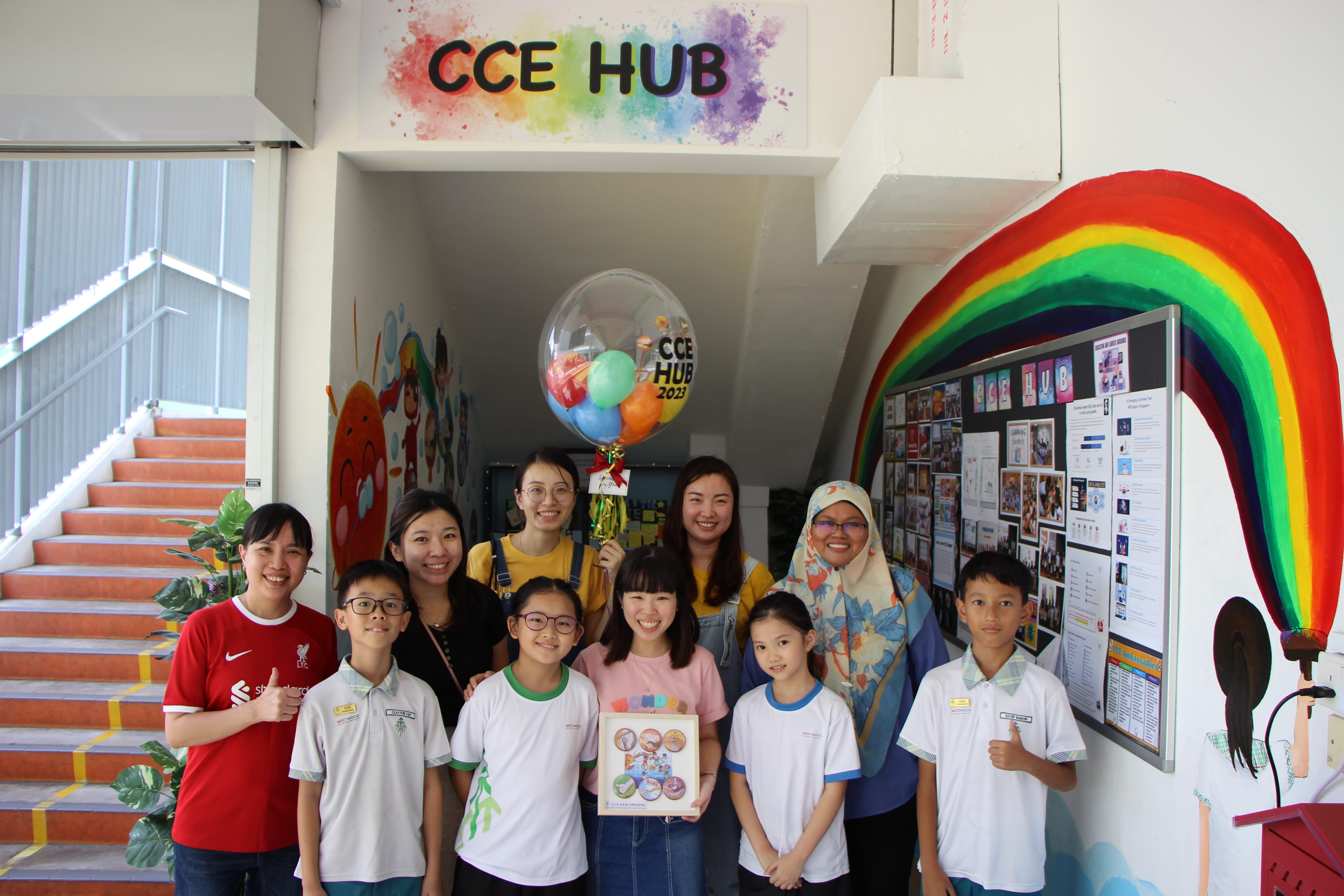 Group of adults and children pose under CCE HUB sign, beside rainbow wall mural and photo bulletin board.